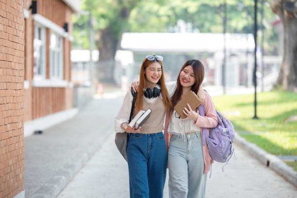 Two female Asian American friends are walking together on their school campus. They are both holding books in their hands with their backpacks on.