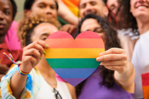 A group of LGBTQ people are standing together as they hold a heart shaped pride flag.