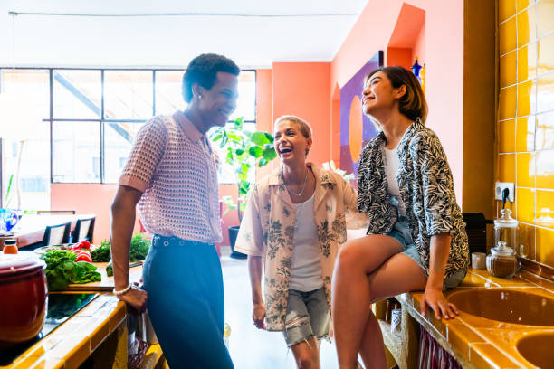 A group of 3 LGBTQ friends are hanging out together in their kitchen. One of females is sitting on the countertop as her 2 friends surround her. They are all laughing together.