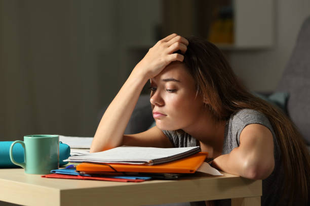 A young teenage girl is sitting in her room on the floor. She is leaning on the table with her notebook open on the desk. She has her hand on her head.