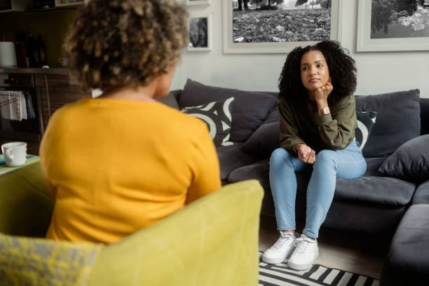 A young African American woman is sitting across from her therapist. She had her hand on her chin as she leans on her knees.