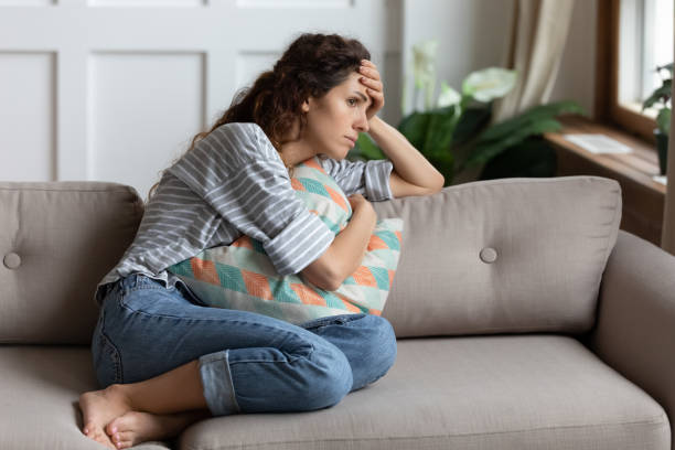 A young woman is sitting on the couch in her living room. She has her legs up on the couch and is holding a pillow tightly on her chest. She has her hand on her forehead.