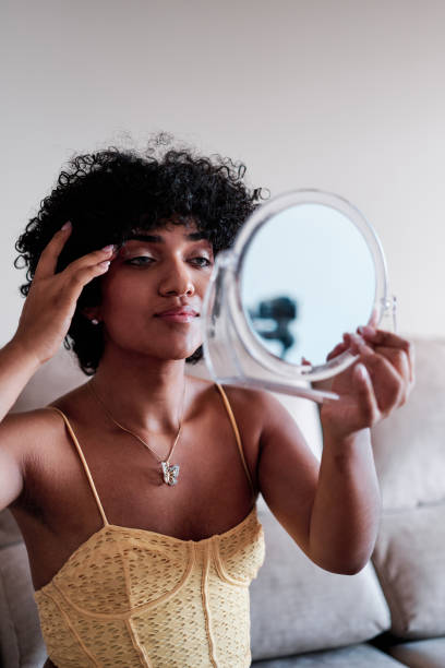 A young LGBTQ woman is holding up a mirror as she looks at herself. She is touching her face.