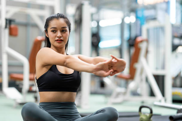 A young Asian American woman is working out at the gym. She is sitting on the floor as she stretches her arms.