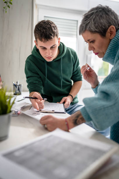 A son and mother are leaning on a table together as they work on an assignment. The son is holding a pen in his hand as he looks down at his paper.