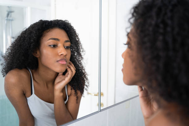 A young African American woman is looking at herself in the mirror in her bathroom. She is leaning into the mirror as she examines her face.