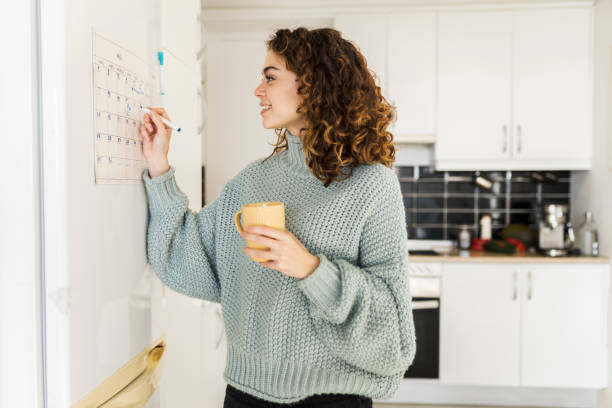 a young woman is standing in her home in the kitchen. She is holding a cup of coffee in her hand as she writes on her calendar that is mounted on the fridge.