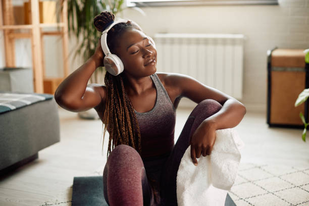 A young African American woman is sitting on a yoga mat in her living room. She is wearing athletic clothing and has headphones on. She is holding a white towel as she touches the back of her head with her eyes closed.