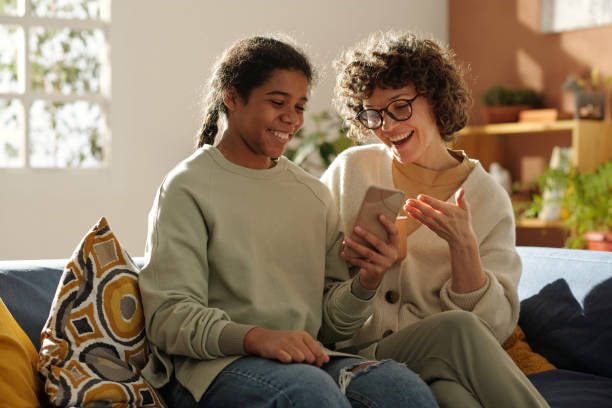 A young mother and her adopted African American daughter are sitting together on the couch of their living room. They are both smiling at the phone that is in the daughters hand.