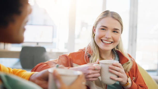 A young woman is sitting with her friend on the couch. They are both holding cups of coffee in their hands as they smile.