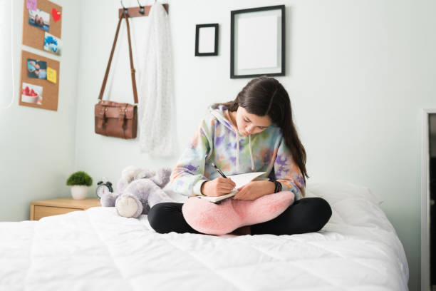 A young female student is sitting on her bed in her room. She is writing in her journal that is on her lap on a pink pillow.