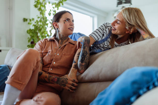 A young lesbian couple is sitting together on their couch. One of the females is laying down while the other sits down.