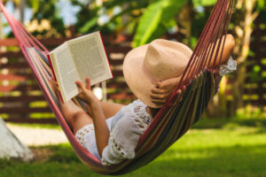 A young woman is laying down outside on a hammock. She is wearing a hat as she reads a book.