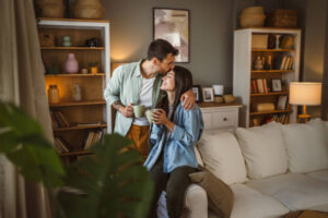 A young couple is sitting in their home on the arm of their couch. The husband is hugging his wife and giving her a kiss on her forehead. They are both holding cups of coffee in their hands.