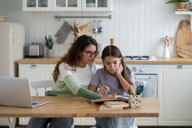 A mother and daughter are sitting on the dining table in their kitchen. The mother is helping her daughter with her homework. There is an open laptop and books in front of them.