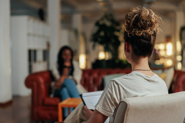 A young woman is sitting across from her therapist in a therapy office. The therapist has a notepad on her lap.