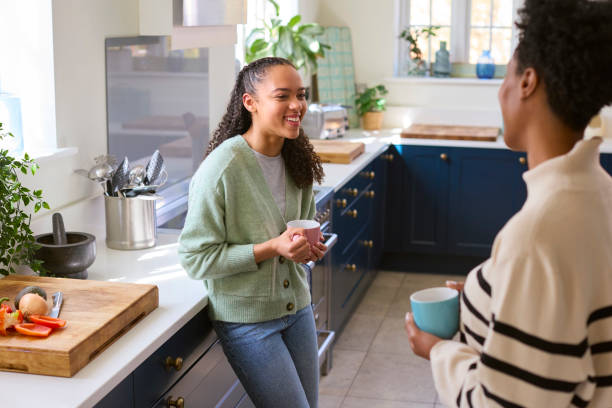 A young African American teenage girl is standing across from her mom in the kitchen. They are both holding coffee cups in their hands as they talk to one another happily.