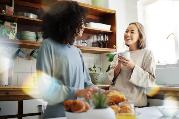 A same-sex couple is standing in a kitchen together. The African American woman is holding a piece of bread in her hand, as her partner stands beside her with a cup of coffee in her hand.