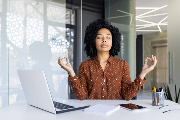 A young African American business woman is sitting in her office with her laptop open in front of her. She has her eyes closed and her hands up beside her as she meditates.