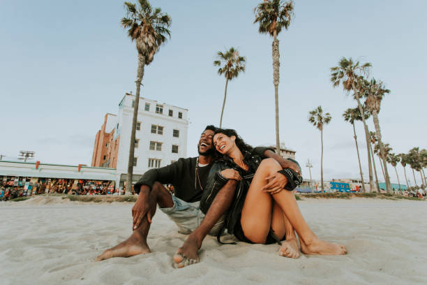 A young biracial couple is sitting on the sand together at the beach. They are leaning into each other as they laugh.