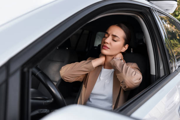 A young woman is sitting in the driver seat of her car with her window down. She has her hands on her neck to relieve her neck pain.