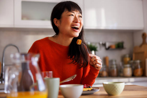 An Asian American woman is sitting in her kitchen as she eats breakfast. She has her fork towards her mouth as she laughs.