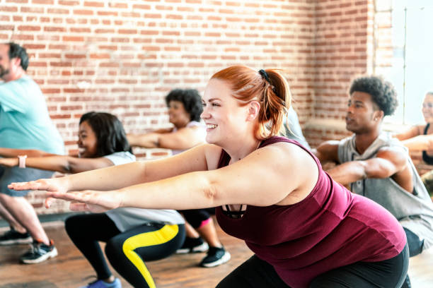 A young woman is in a exercising class as she does a squat. Beside her are multiple people doing the same pose as her.