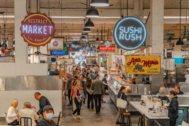 This image is a market place filled with people. On the left there is a light-up sign that says "District Market Los Angeles" and on the right there is a blue light-up sign that says "Sushi Rush".