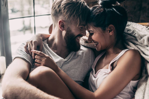 A young couple is snuggling together with a blanket on the couch. Their foreheads are touching each other as they smile and laugh.