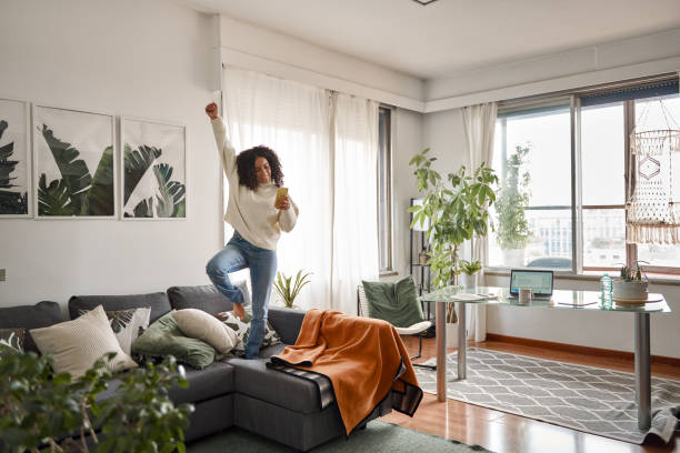 A young African American woman is standing on her couch celebrating. She is raising her hand in celebration as she looks at her phone in her hand.