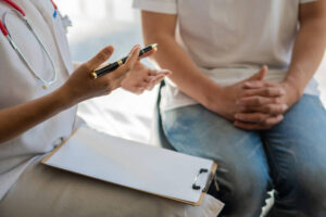 A woman is sitting beside her doctor with her hands on her lap. The doctor has a notepad on her lap and a pen in her hand.