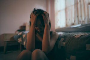 An Asian American woman is sitting on the floor of her bedroom with dim lights. She has her hands on her head as she looks down.