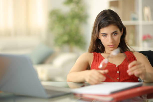 A young woman is sitting at her desk in her home. She has her laptop open in front of her and a notebook on the table. She is fidgeting with her glasses.