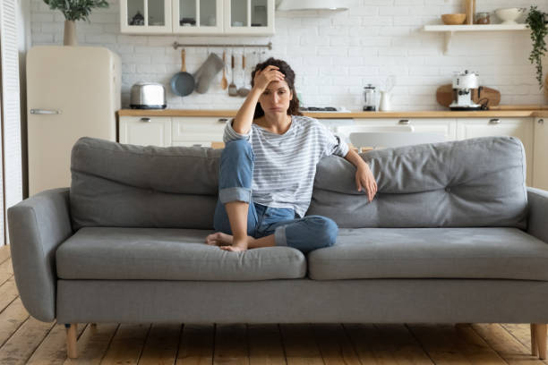 A young woman is sitting on her couch in her home. She has her legs up on the couch as she puts her hand on her forehead.