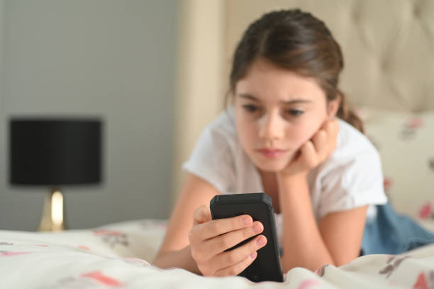 A young teenage girl is laying on her bed in her room. She is looking at her phone with a concerned face.