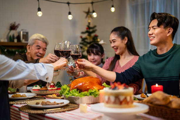 An Asian American family is sitting together at the dinner table for Thanksgiving. The adults are doing a cheers with red wine.