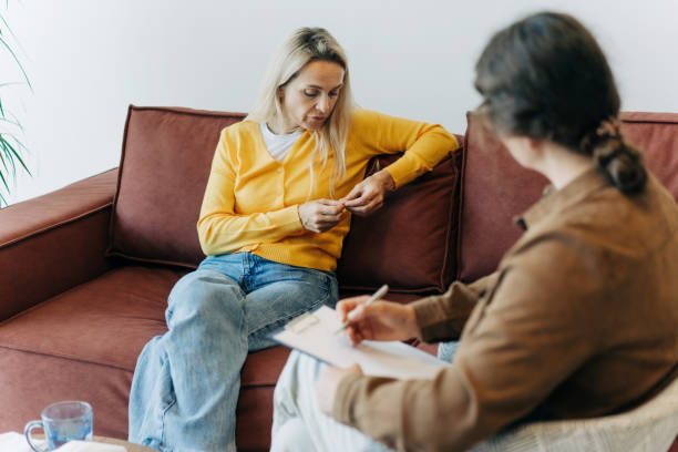 A young woman is sitting across from her therapist on the couch. The woman is playing with her hands. The therapist has a notepad on her lap.