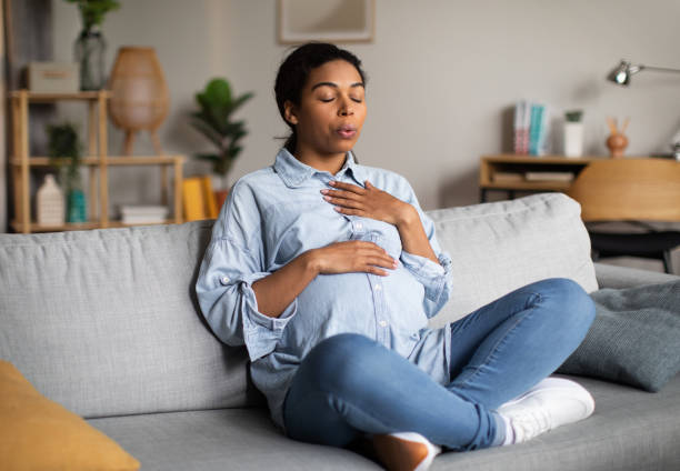 A pregnant African American woman is sitting with her legs crossed on her couch in her living room. She has one hand on her belly and one hand on her chest as she breathes.