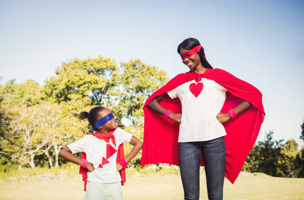 An African American mother and daughter are dressed up for Halloween. They are both wearing a red cape, a superhero mask, and a t-shirt with a red symbol on their chest.