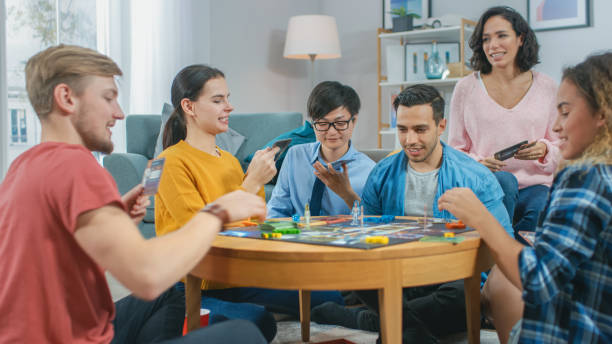 A group of 5 teenagers are sitting on the floor and playing a board game together. One of their mothers is sitting behind them as she participates in the game.