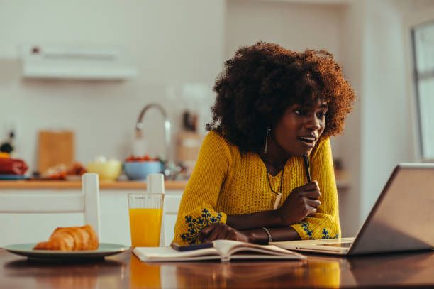 A young African American woman is sitting at her dining table in her home. In front of her is her open laptop and a pen in her hand.