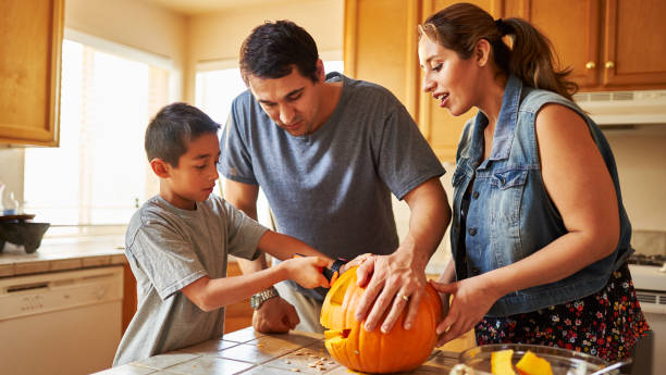 A Hispanic family of three are in their kitchen carving a pumpkin together. The parents are helping the young boy as she carves a face onto the pumpkin.