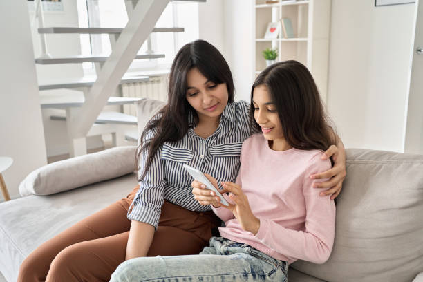 A young woman is sitting beside her young teenage daughter in their living room. The daughter is using her phone as they both look at the screen.