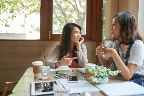 Two young Asian American females are sitting at a cafe having coffee together. They are both smiling as they speak.