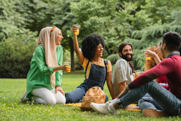 A group of 5 teenage friends are having a picnic outdoors. They are all sitting on the grass as they drink a juice and laugh together.