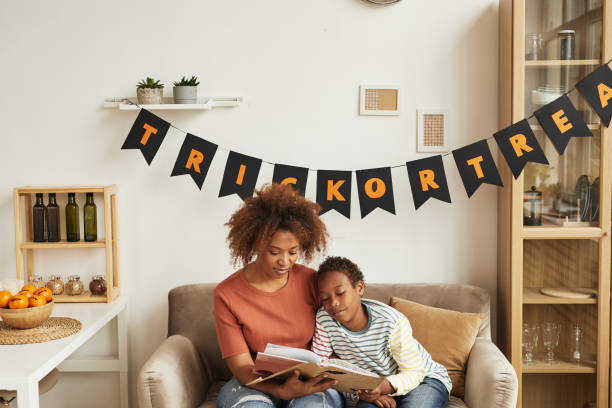 An African American mother and son are sitting in their living room on the couch, reading a book together. Behind them is a banner on the wall that says "trick or treat".