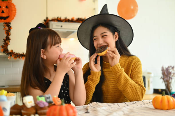 A young Asian American mother and her daughter are wearing witches hats as they both eat donuts. The room is decorated with Halloween decorations.