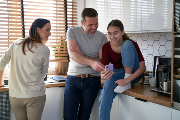 A family of three is spending time together in their kitchen. The parents are looking at their daughters phone as they laugh and smile.