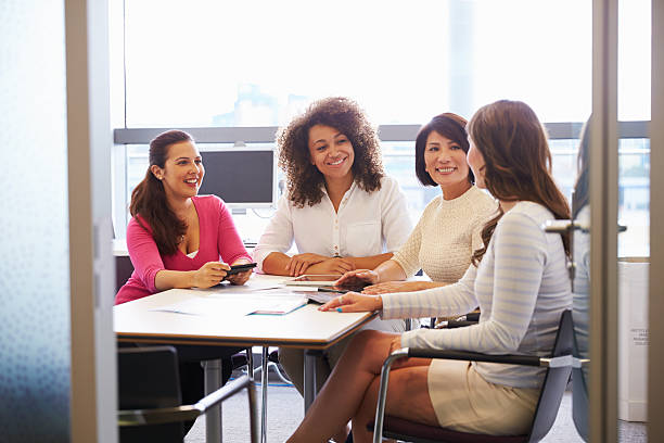 A group of four business women are sitting at a table in their office having a meeting. They are are smiling.