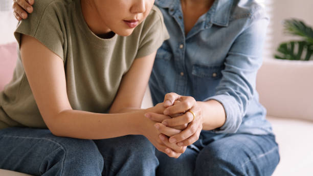 A mother and her teenage daughter are sitting beside each other on the couch. The teen girl is hunched over with her hands on her knees as her mother puts her arm around her.